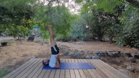 תנוחת סרוונגאסנה – עמידת כתפיים מרגיעה וממריצה גבר בתנוחת עמידת כתפיים, שוכב על גבו עם רגליים מורמות ישרות לשמיים, מוקף בטבע ירוק