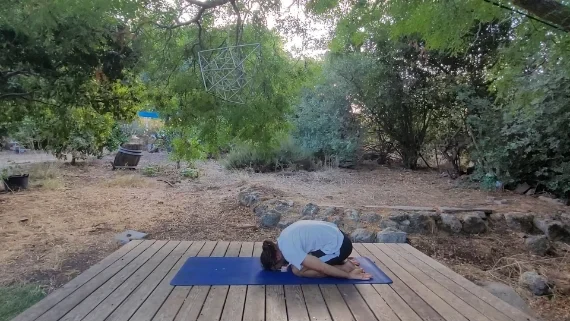 תנוחת הילד – מנוחה, שחרור והרפיה גבר בתנוחת הילד, ישוב על העקבים וראשו מונח קדימה על המזרן, מוקף בטבע ירוק